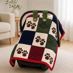 May include: A colorful, square-patterned baby blanket with paw prints, draped over a car seat. The blanket features squares of white, navy, burgundy, and green, with brown paw prints. The edges are trimmed with red yarn.