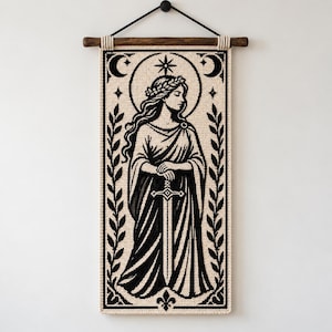 Può includere: Un arazzo rettangolare intrecciato color crema e nero raffigurante una figura che indossa una corona d'alloro e una veste drappeggiata, mentre impugna una spada. Il design include una stella, lune crescenti e motivi a foglie, appeso a un bastone di legno scuro con un cordino nero.