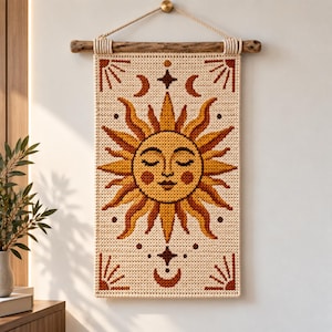 Può includere: Un arazzo rettangolare con un volto di sole all'uncinetto con occhi chiusi e guance rosate, circondato da raggi in tonalità di giallo e marrone. Il design include mezzelune e motivi a stella su uno sfondo color crema, appeso a un ramo.
