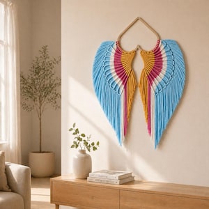 May include: A vibrant macrame wall hanging shaped like wings, with blue, pink, yellow, and white fringe. The artwork is mounted on a wooden frame and hangs against a neutral wall. The piece adds a pop of colour.
