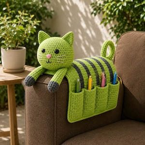 May include: A handmade, green crochet cat-shaped armrest organizer with gray stripes and four pen holders. The cat has a pink nose, black eyes, and gray paws. Pens of various colors are in the pockets. The organizer is placed on a brown sofa.