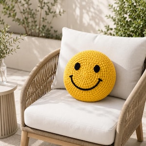 May include: A yellow, circular, crocheted cushion featuring a smiley face design. The cushion has black eyes and a black curved mouth. It sits on a white cushion of a beige woven chair.