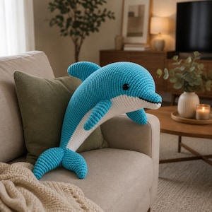 Könnte beinhalten: Ein großer, handgehäkelter Delfin-Kissen in Blau und Weiß. Der Delfin hat ein lächelndes Gesicht mit schwarzen Augen und einen detaillierten Schwanz und Flossen. Das Kissen liegt auf einem beigen Sofa, was auf eine nautische Dekoration hindeutet.