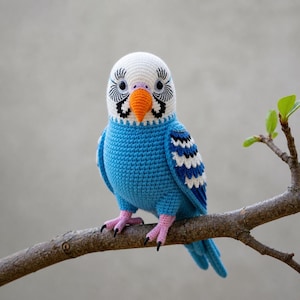 May include: A handmade crochet parakeet in shades of blue, white, and orange, perched on a brown branch. The bird features detailed stitching, including black and white eye accents and pink feet. The background is a soft gray.