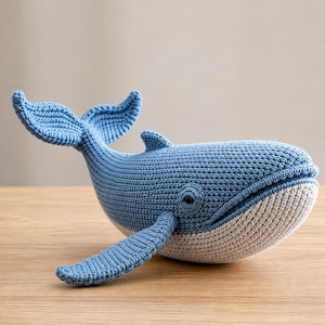 May include: A handmade blue and white crochet whale toy. The whale features a detailed design with fins, a tail, and a rounded body. The toy is made of soft yarn and is suitable for children.