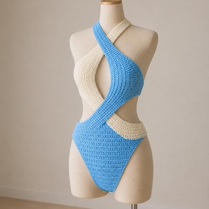 May include: A handmade, two-tone crochet swimsuit in light blue and cream. The halter-style top crosses over the chest and back, with a high-cut bottom. The swimsuit is displayed on a mannequin.