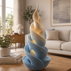 May include: A unique, handcrafted crochet lamp in a spiral design. The lamp transitions from a light beige at the top to a light blue at the base. The lamp has a warm glow, and is placed on a wooden table.