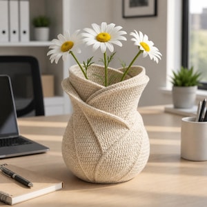 Peut inclure: Un vase beige en crochet avec un design plié unique, contenant trois marguerites blanches à cœur jaune. Le vase est posé sur un bureau en bois, présentant une composition florale artisanale. Le vase mesure environ 20 cm de haut.