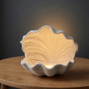 Peut inclure: Lampe en forme de coquillage blanc avec une surface texturée. La lampe est ouverte, révélant une lumière chaude et brillante à l'intérieur. Les bords de la coquille ont un design ondulé. La lampe est posée sur une table en bois.