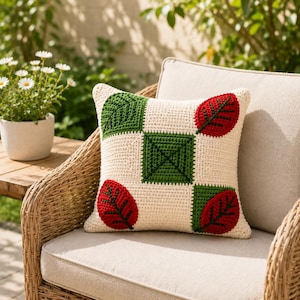 May include: Cream-colored crochet pillow with a leaf design. The pillow features four squares with green and red leaf patterns. The pillow is placed on a wicker chair with a beige cushion, with a potted plant in the background.