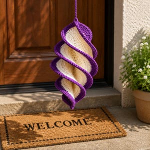 May include: A hanging, spiral-shaped crochet decoration with a purple border and cream-colored center. The decorative piece is suspended by a purple cord. A welcome mat is visible below.