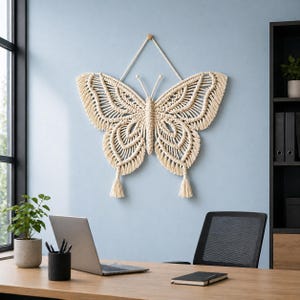 Può includere: Una grande decorazione da parete a farfalla in macramè color crema. La farfalla presenta intricati motivi di nodi e nappe decorative. È sospesa a un tassello di legno e a una corda contro un muro azzurro.