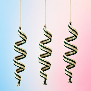 May include: Three spiral-shaped crochet decorations. Each is cream-colored with a dark green stripe, suspended from a cream-colored string. The crochet work is visible, and each spiral ends in a small tassel. The background fades from blue to pink.