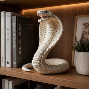 May include: A cream-coloured, crocheted cobra sculpture with an open mouth, displayed on a wooden shelf. The snake is coiled and detailed, with visible stitching. Books and a framed picture are in the background, suggesting a home decor theme.