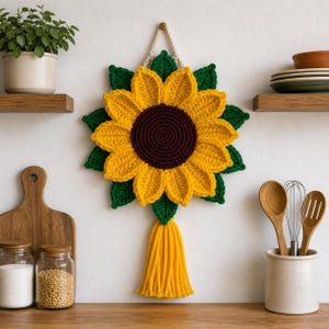 May include: A handmade, yellow crochet sunflower wall hanging with a dark brown center and green leaves. The sunflower is adorned with a yellow tassel and a cream-colored rope for hanging. The sunflower is approximately 12 inches in diameter.