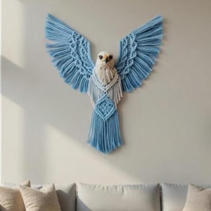 Puede incluir: Un tapiz de macramé que representa un águila con las alas extendidas. El águila presenta un cordón de algodón anudado en azul claro y blanco. Las alas están muy extendidas y el cuerpo tiene un diseño en forma de diamante. El águila está montada en una pared de color neutro.