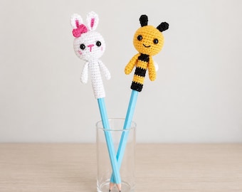 Puede incluir: Dos lápices en un vaso transparente, cada uno coronado con un animal de ganchillo. Uno es un conejito blanco con un lazo rosa, el otro una abeja amarilla y negra. Los lápices son de color azul claro.