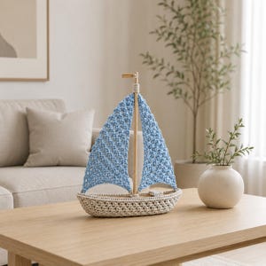 Può includere: Una barca a vela all'uncinetto fatta a mano con una vela azzurra e un albero in legno naturale. La barca è posta su una base all'uncinetto beige. L'oggetto nautico decorativo è esposto su un tavolo di legno.