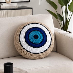 May include: A round, beige crochet pillow with a blue and white eye design. The eye features concentric circles of dark blue, light blue, and white, centered on a turquoise blue circle. The pillow is placed on a cream-colored sofa.