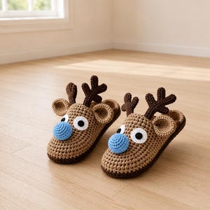 May include: A pair of handmade crochet reindeer slippers. The slippers are brown with dark brown antlers, large white-rimmed eyes, and blue noses. The slippers are designed to look like a reindeer's head.