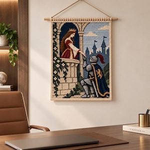 Op de afbeelding: Een rechthoekige wandhanger met een kruissteekontwerp van een koningin in een rode jurk op een stenen balkon en een ridder in blauw harnas die voor haar knielt. De scène bevat een kasteel op de achtergrond en klimmende witte bloemen.