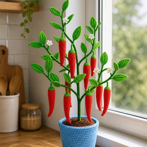 Op de afbeelding: Een gehaakte chilipeperplant in een blauwe pot. De plant heeft felrode chilipepers, groene bladeren en kleine witte bloemen. De pot staat op een houten oppervlak, met een raam en keukenartikelen op de achtergrond.