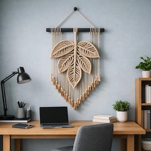Könnte beinhalten: Beige Makramee-Wandbehang in Blattform, aufgehängt an einer schwarzen Holzstange. Das Blatt hat detaillierte Adern, und der Boden hat Quasten. Die Wanddekoration wird über einem Holzschreibtisch mit Laptop und Lampe ausgestellt.