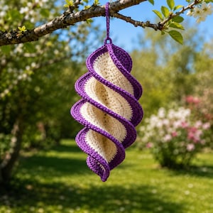 May include: A handmade, spiral-shaped crochet hanging decoration. The piece features a cream-colored center with a vibrant purple border and spiral accents. It is suspended from a tree branch against a blurred background of greenery and flowers.