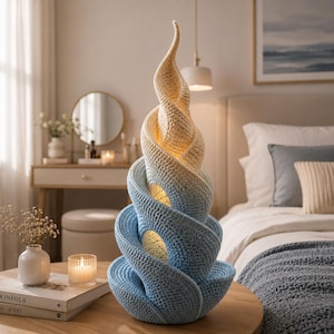 Puede incluir: Lámpara de ganchillo hecha a mano con un diseño en espiral. La lámpara pasa de una base azul claro a una parte superior color crema. La lámpara tiene un aspecto texturizado y emite un brillo cálido. Es una pieza decorativa.