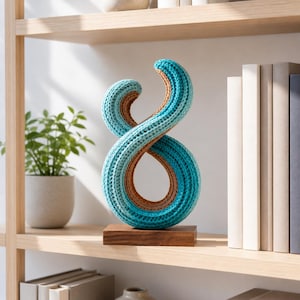 May include: A decorative sculpture in the shape of an infinity symbol, crafted from knitted material. The sculpture features a combination of teal, brown, and light blue colors. It is mounted on a small, rectangular wooden base, and is displayed on a wooden shelf.