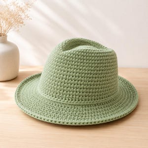 Op de afbeelding: Een lichtgroene gehaakte fedora hoed met een brede rand. De hoed is gemaakt van een getextureerd, geweven materiaal en staat op een lichtgekleurd houten oppervlak. Een witte vaas met gedroogde bloemen staat op de achtergrond.