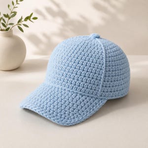 Può includere: Cappellino da baseball all'uncinetto azzurro. Il cappellino presenta una corona arrotondata, una visiera curva e un piccolo dettaglio a bottone sulla parte superiore. La trama del punto all'uncinetto è visibile su tutto il cappellino.
