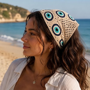 May include: A beige crochet headscarf with a repeating pattern of blue and navy blue evil eye designs. The headscarf is square-shaped and worn on the head. The background shows a beach and ocean.