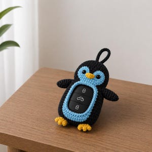 Puede incluir: Una funda para llavero de pingüino de ganchillo negro y azul con detalles amarillos. La funda para llavero tiene un lazo para sujetarlo a un llavero y presenta un mando a distancia para coche. El pingüino tiene la barriga azul y las patas amarillas.