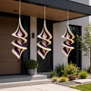 Könnte beinhalten: Drei hängende, spiralförmige Häkelornamente. Jedes Ornament hat abwechselnd cremefarbene und violette Streifen, die an einem dünnen, durchsichtigen Faden aufgehängt sind. Die dekorativen Stücke werden im Freien ausgestellt und verleihen dem Äußeren einen handgefertigten Charme.