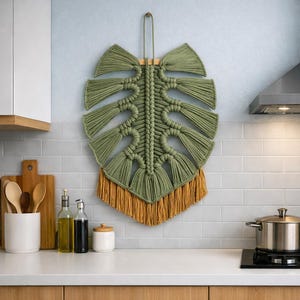 May include: A large, green macramé leaf wall hanging with a mustard-coloured fringe. The leaf design is mounted on a wooden dowel and hangs against a light blue tiled wall. The macramé is made of thick, woven cord.