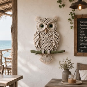 Könnte beinhalten: Ein Wandbehang aus Makramee in Form einer Eule mit grünen Augen und einem Holzschnabel, der auf einem grünen Ast sitzt. Die Eule ist aus beigem Seil gefertigt und hängt an einer weißen Wand. Im Hintergrund ein Café mit einer Tafel.