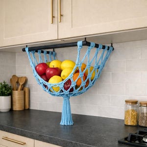 May include: A light blue macrame fruit hammock filled with red apples, yellow lemons, and orange oranges. The hammock is suspended from a black rod and hangs over a kitchen countertop. A blue tassel hangs from the bottom.