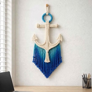 Könnte beinhalten: Wandbehang im maritimen Stil in Form eines Ankers. Der Anker besteht aus cremefarbenem Seil mit einem blauen Ombre-Fransen. Ein blauer Seilkreis und ein Holzdübel befinden sich oben zum Aufhängen. Der Anker wird an einer weißen Wand gezeigt.