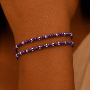 Op de afbeelding: Een dubbelstrengs armband met kleine, ronde, dieppaarse kralen afgewisseld met gepolijste, reflecterende zilverkleurige metalen kralen. Het ontwerp bestaat uit twee parallelle rijen kralen die in een consistent patroon zijn geregen.