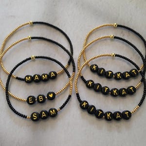 May include: Six circular beaded bracelets featuring small black and gold beads. Three bracelets on the left display the text MAMA, E B with a heart symbol, and SAM. Three bracelets on the right display the text KIT KAT in gold lettering.