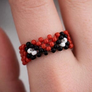 May include: A beaded ring featuring a woven pattern of small, round beads in vibrant red, solid black, and bright white. The design consists of a red base with alternating black and white bead clusters arranged in a repeating, textured grid structure.