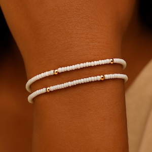 Puede incluir: Dos pulseras de cuentas blancas con detalles dorados. Las pulseras se llevan en la muñeca. Las pequeñas cuentas espaciadas tienen una cuenta dorada a intervalos. Joyería delicada y elegante.