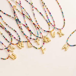 May include: A collection of necklaces featuring multi-coloured seed beads arranged in a vibrant pattern. Each necklace includes a single, polished gold-tone metal letter charm, specifically the letters M, U, W, L, T, Y, Q, V, X, and H.