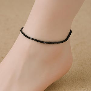 Può includere: Una semplice cavigliera composta da un unico filo di piccole perline nere opache e uniformi. Il gioiello circolare si indossa attorno alla caviglia e presenta un design continuo ed essenziale, privo di chiusure visibili o altri elementi decorativi.