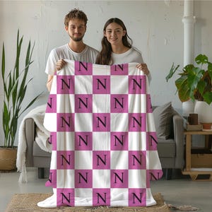 May include: A soft, plush throw blanket featuring a repeating checkerboard pattern of white and magenta squares. Each magenta square is printed with a bold, black capital letter N in a serif font, creating a consistent, grid-like design.