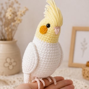 Puede incluir: Un pájaro cacatúa hecho a mano con ganchillo, con cresta amarilla, cabeza y mejillas naranjas. El cuerpo del pájaro es blanco, con ojos negros y patas rosas. El pájaro está posado en una mano.