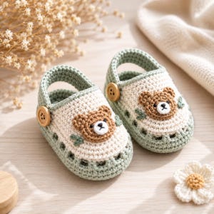 Puede incluir: Un par de zapatos de ganchillo para bebé en color verde salvia y crema, con una aplicación de cara de oso marrón en la parte delantera, un pequeño botón de madera en la tira lateral y un diseño recortado en la base, tejido con punto texturizado.