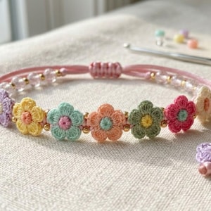 Könnte beinhalten: Ein handgefertigtes Häkelarmband mit bunten Blumenmustern. Das Armband hat eine rosa Kordel mit kleinen goldenen Perlen und einen Schiebeknotenverschluss. Die Blumen sind in Gelb-, Grün-, Rosa- und Orangetönen gehalten.