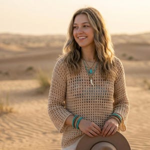 May include: A beige, open-weave sweater with long sleeves. The sweater is paired with turquoise and brown bracelets and necklaces. A brown hat is held in the hands. The background is a desert landscape.